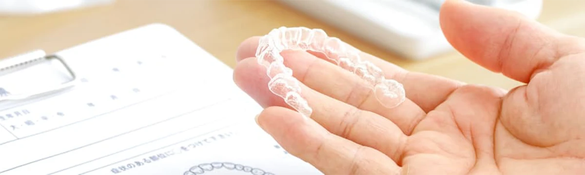 手のひらに透明な歯列矯正用マウスピース（アライナー）を持っている様子。デスク上に資料も見える。