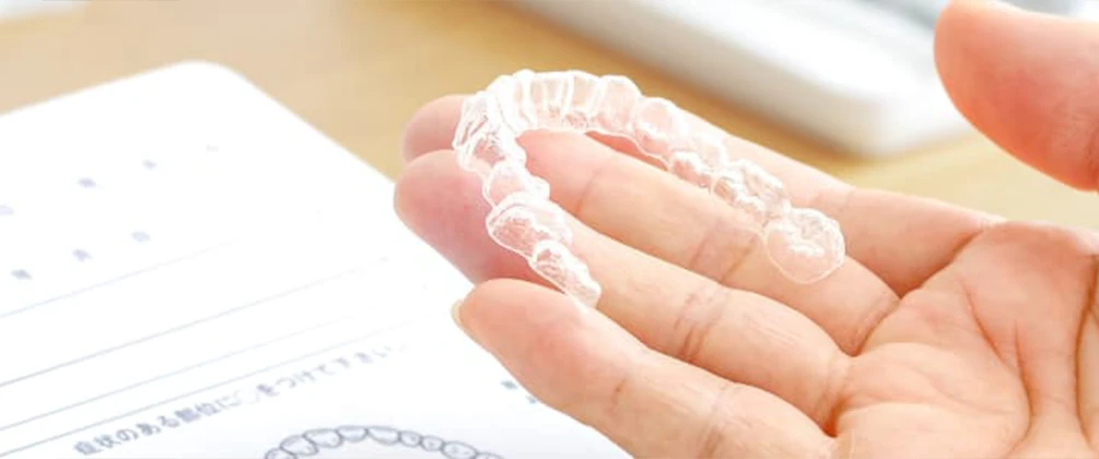 手のひらに透明な歯列矯正用マウスピース（アライナー）を持っている様子。デスク上に資料も見える。